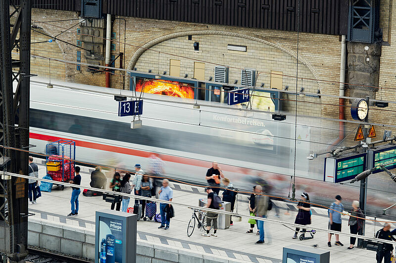 Menschen am Bahnhof, im Hintergrund einfahrender Zug