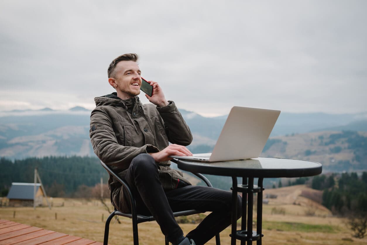 Mann sitzt am in den Bergen draußen am Tisch mit Laptop und telefoniert