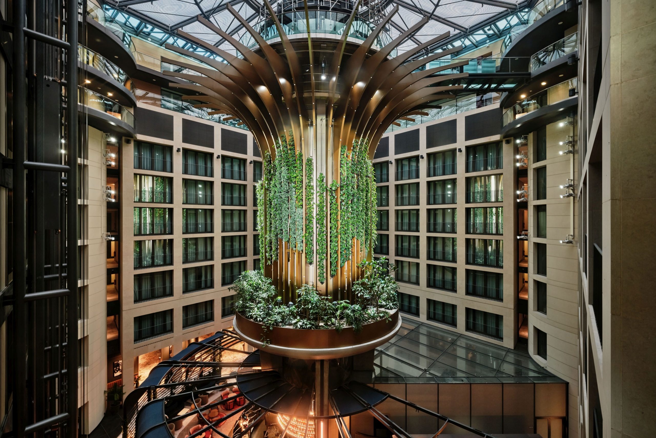 Modernes Hotelatrium mit zentraler Pflanzeninstallation, geschwungener Deckenstruktur aus Holz und Glas, umgeben von mehrgeschossigen Zimmertrakten.