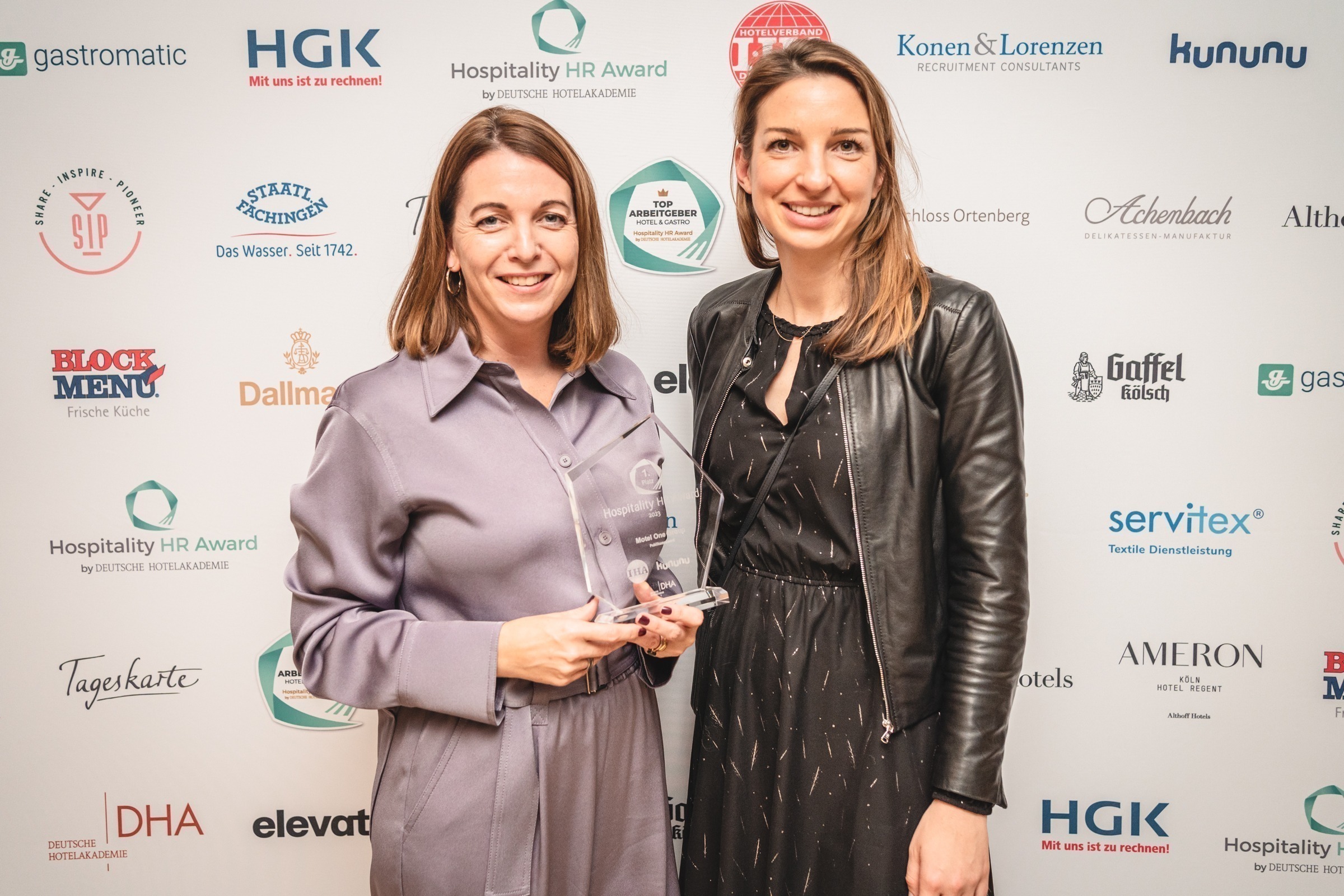 Zwei Personen bei Preisverleihung des Hospitality HR Awards, eine im lilafarbenen Outfit mit Auszeichnung, eine im schwarzen Outfit mit Lederjacke.