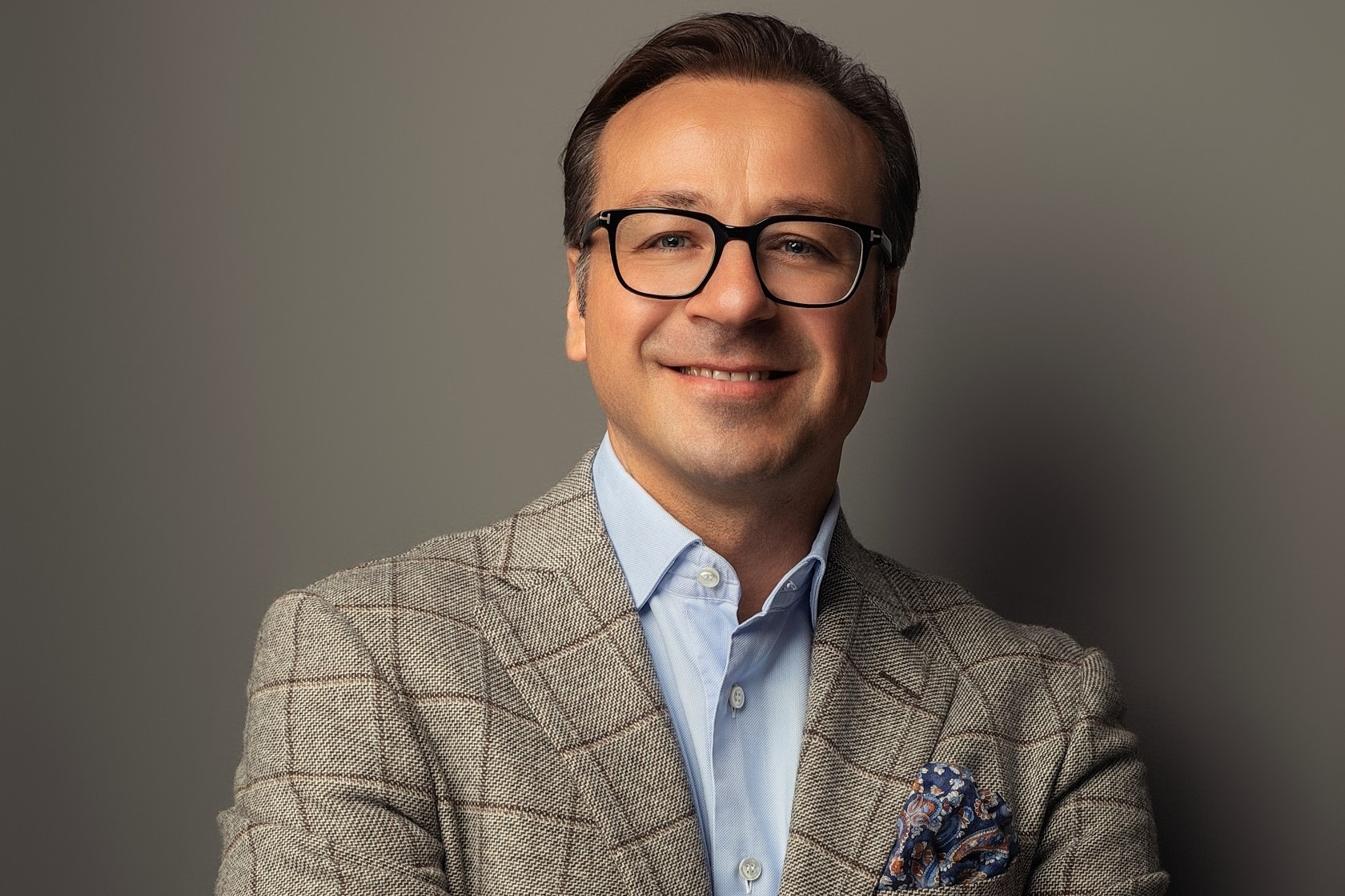 Professionelles Porträt eines lächelnden Mannes mit Brille in grau kariertem Anzug, hellblauem Hemd und gemustertem Einstecktuch