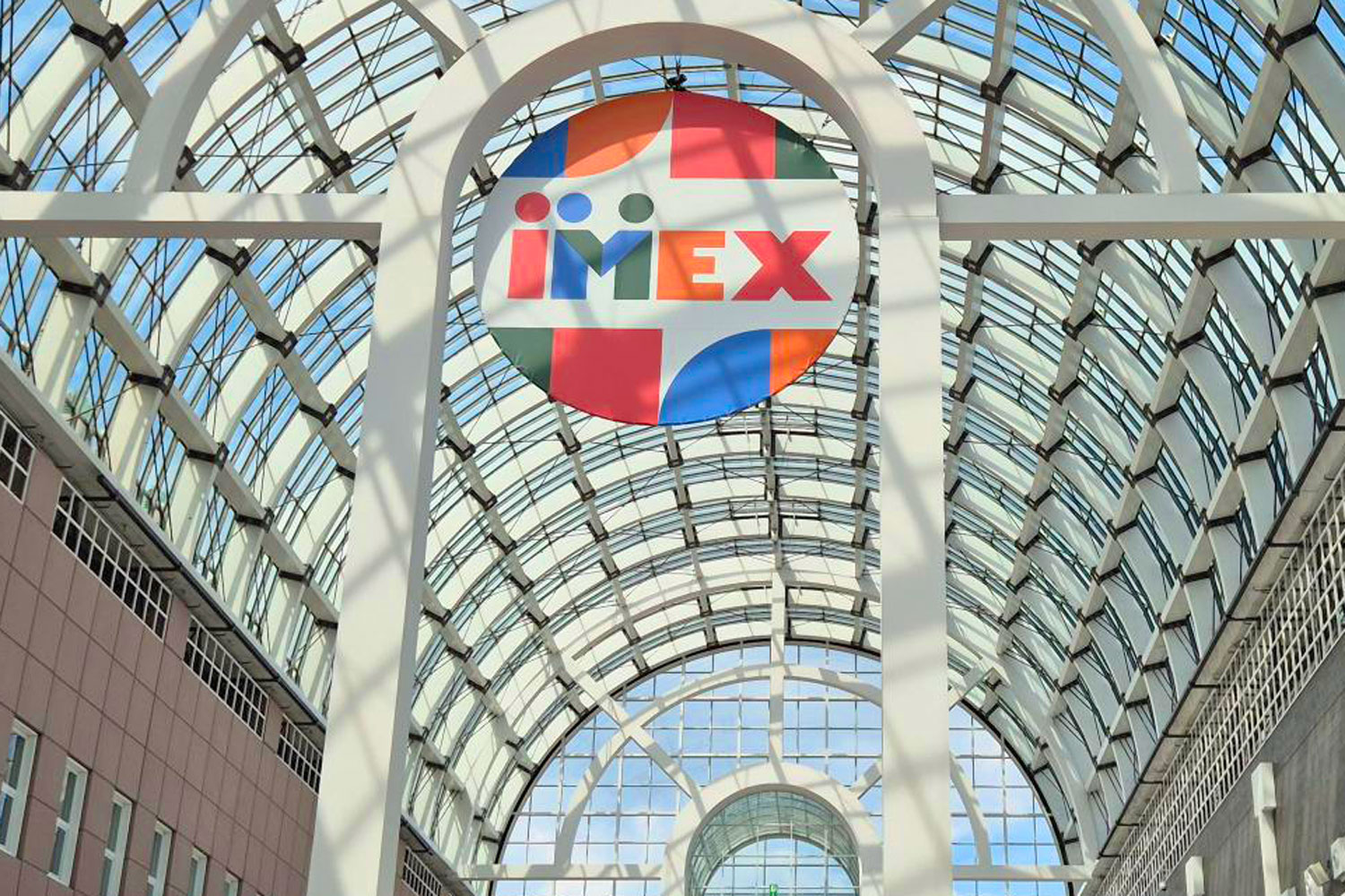 Glasdach einer Messehalle mit großem bunten IMEX-Logo im Zentrum, umgeben von weißen Stahlträgern und blauem Himmel.