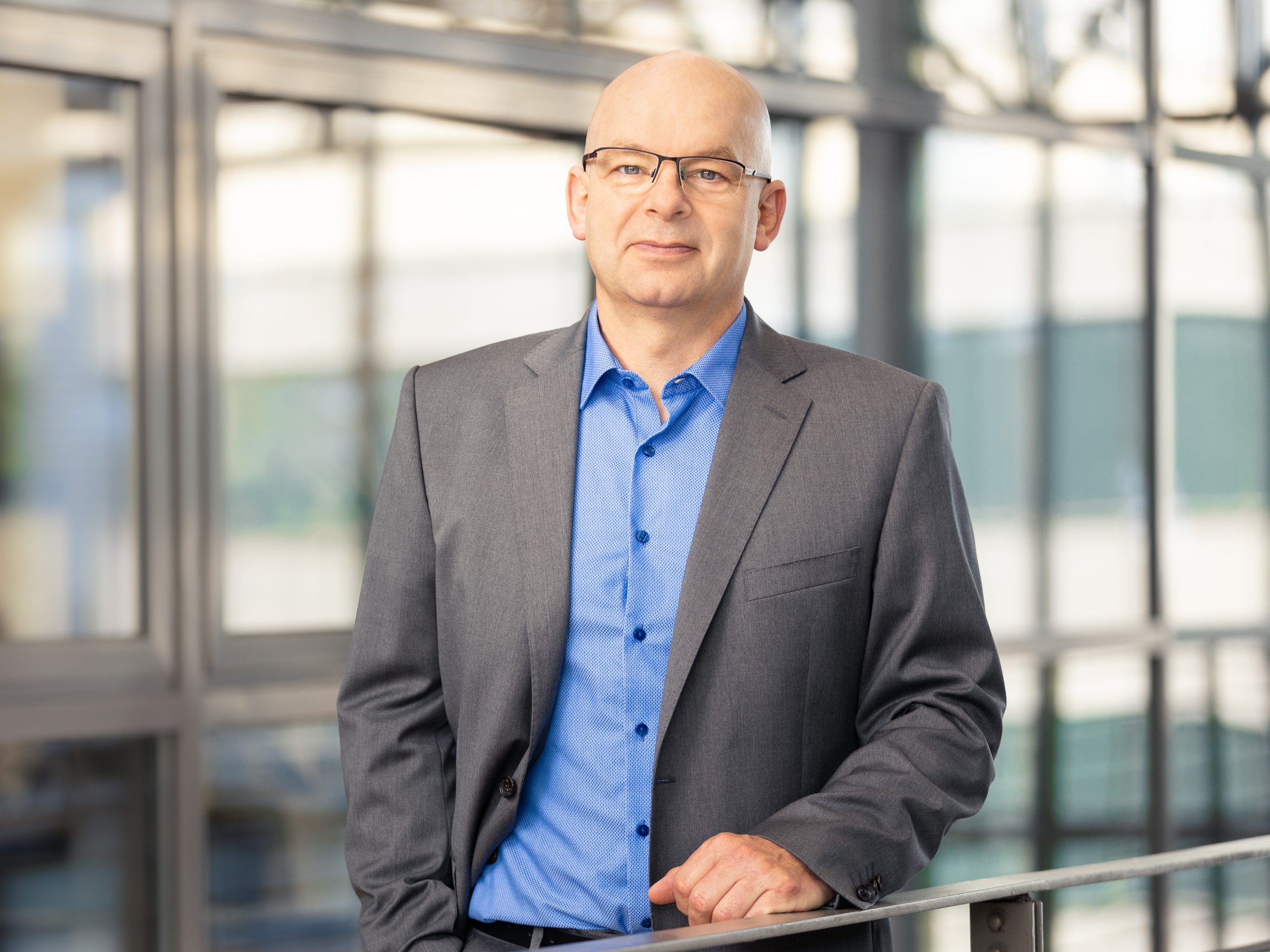 Ein glatzköpfiger Mann mittleren Alters mit Brille steht in einem modernen Bürogebäude. Er trägt ein graues Sakko über einem blauen Hemd und blickt selbstbewusst in die Kamera. Im Hintergrund sind große Fensterfronten mit Glasrahmen und unscharfe Außenansichten zu sehen