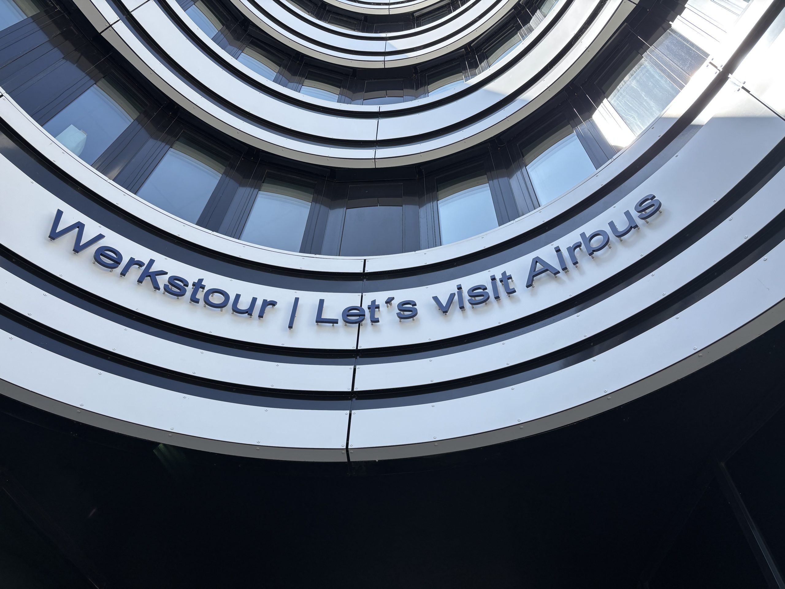 Gebäude mit gebogener Fassade und reflektierenden Fenstern. An der Außenwand steht in blauen Buchstaben: „Werkstour | Let’s visit Airbus“.